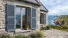 Volets battants PVC persiennés gris-bleu sur une maison en granit face à l'océan en Bretagne, phare au loin